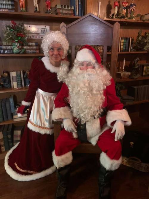 A woman in a vintage dress stands beside Santa Claus, who is seated in a large chair, surrounded by books and decorations.