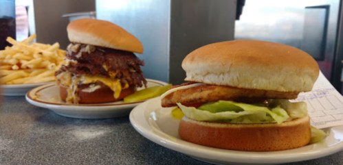 Two burgers on plates: one with grilled chicken and lettuce, the other stacked with beef and cheese, alongside fries.