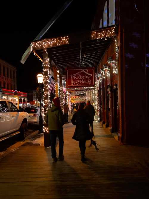 Two people walk along a festive, lit-up street at night, passing by a café adorned with holiday lights.