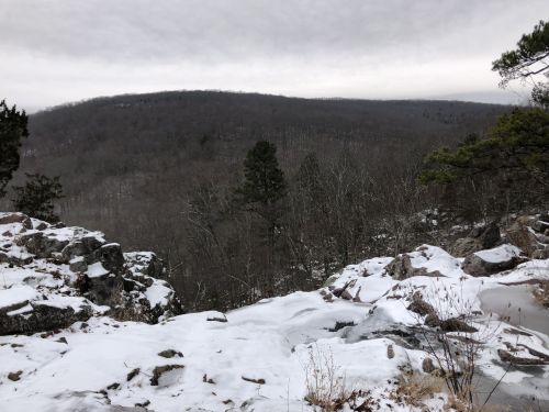 A snowy landscape with rocky outcrops and a view of a distant, forested hill under a cloudy sky.