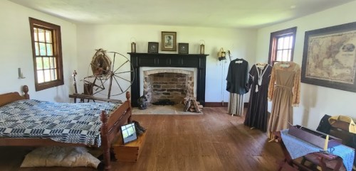 A historic room featuring a bed, spinning wheel, fireplace, and period clothing on display.