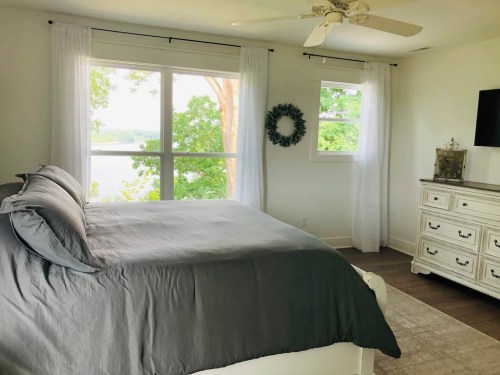 Bright bedroom with a gray bedspread, large windows, and a view of greenery outside. White dresser and light curtains.