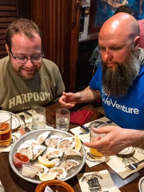 Two men sit at a table, excitedly looking at a platter of oysters with lemon and sauces, drinks nearby.