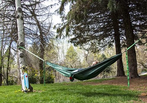 A person relaxes in a green hammock tied between two trees in a grassy area surrounded by nature.