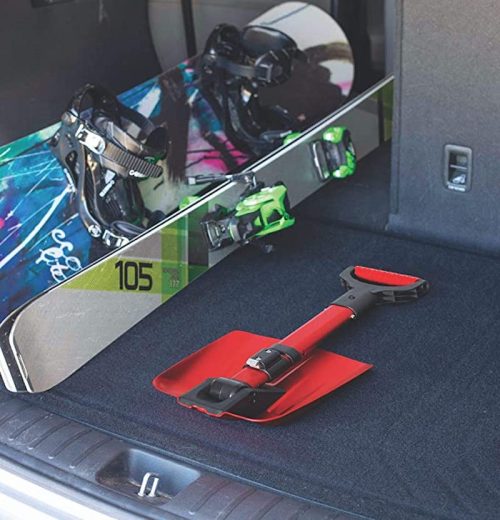 A snowboard and bindings are placed in the trunk of a car, alongside a red shovel on a black carpeted surface.