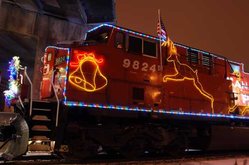 A brightly lit train decorated with colorful Christmas lights and festive designs, parked under a bridge at night.