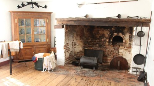 A rustic room featuring a large brick fireplace, wooden furniture, and various kitchen tools.