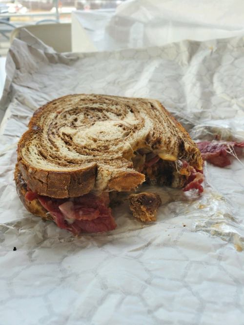 A half-eaten sandwich on crumpled paper, featuring marbled rye bread and layers of meat and cheese.