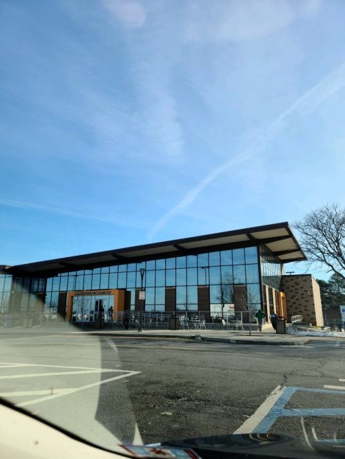 Modern glass building with a flat roof, surrounded by a parking lot and trees under a clear blue sky.