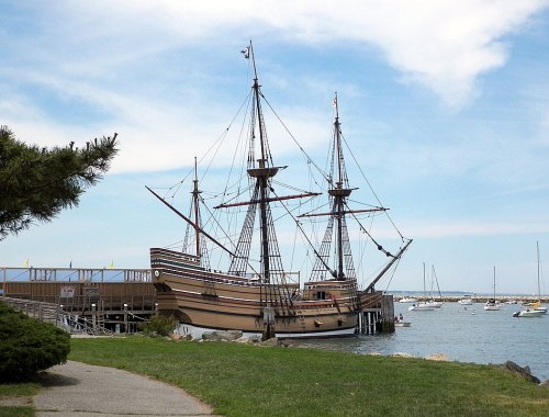 A historic ship docked by the shore, surrounded by water and sailboats under a blue sky.