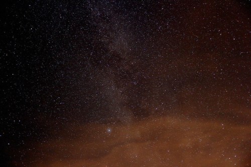 A starry night sky filled with countless stars and a faint Milky Way, with clouds illuminated by a warm glow.