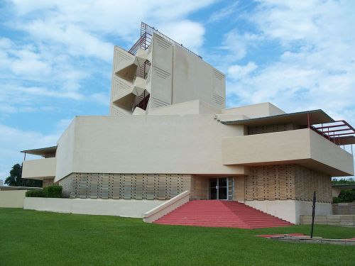 A modernist building with geometric shapes, featuring a light-colored facade and red steps, set against a blue sky.