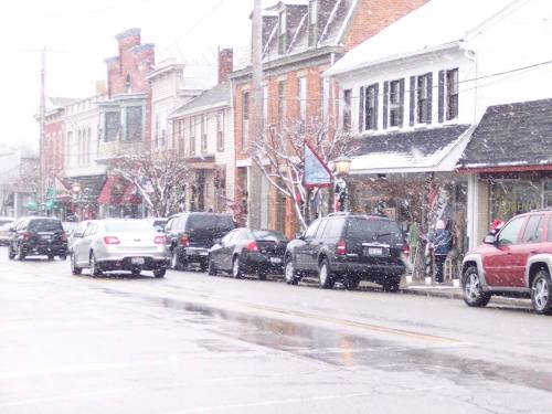 A snowy street scene with parked cars and quaint shops, decorated for the winter season.