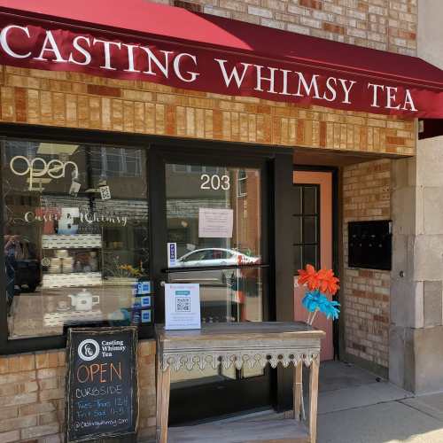 A storefront with a red awning reading "Casting Whimsy Tea," featuring an open sign and a decorative entrance.