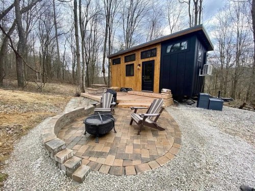 A small modern cabin with a wooden deck, surrounded by trees and a stone patio with chairs and a fire pit.