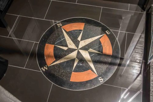 A decorative compass rose embedded in a dark tiled floor, featuring cardinal directions: N, E, S, W.