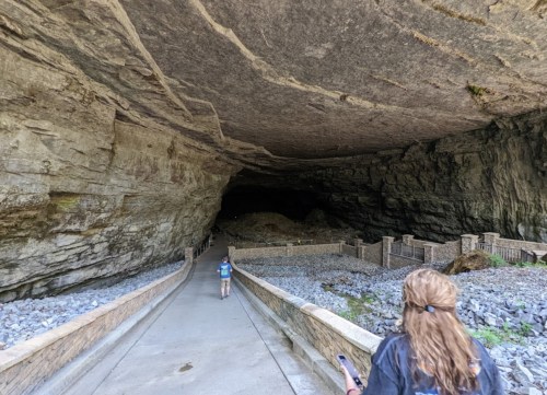 A person walks towards a large cave entrance, with rocky walls and a gravel path leading inside.
