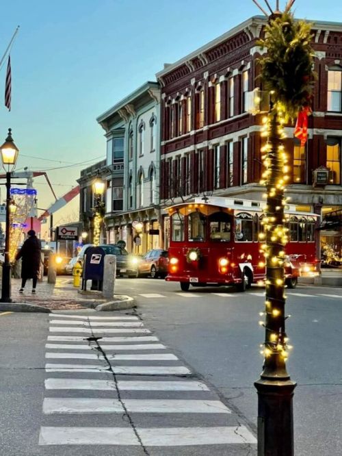 A charming street scene at dusk, featuring a red trolley, festive lights, and historic buildings.