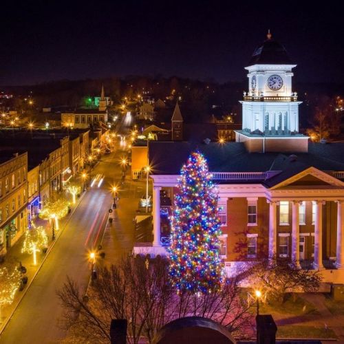 A festive town square at night, featuring a brightly lit Christmas tree and illuminated buildings.