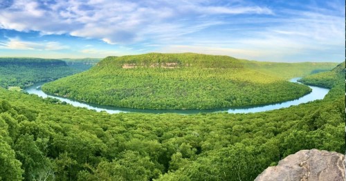A panoramic view of a lush green landscape with a winding river and a prominent hill under a blue sky.
