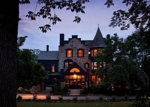 A historic stone mansion with a turret, illuminated windows, surrounded by trees at dusk.