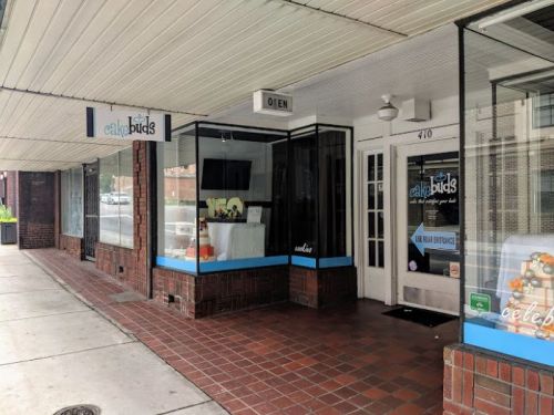 Exterior view of a storefront with large windows, featuring a sign that reads "Cake Buds" and an "Open" sign.