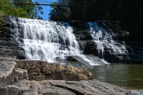 A serene waterfall cascades over rocky ledges into a calm pool, surrounded by lush greenery and trees.
