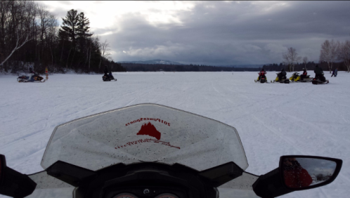 View from a snowmobile on a snowy landscape, with other snowmobiles and trees in the background under a cloudy sky.