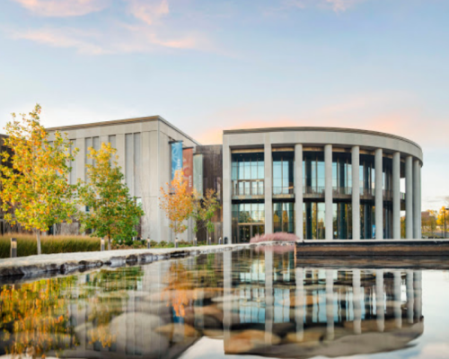 Modern building with large glass windows, surrounded by trees and reflecting in a calm water feature at sunset.