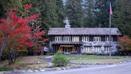 A rustic stone and wood lodge surrounded by trees, with autumn foliage and a flagpole in front. Smoke rises from the chimney.