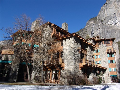 A rustic stone and wood lodge surrounded by snow and mountains under a clear blue sky.