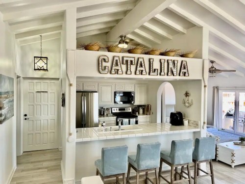 Bright kitchen with white walls, a large "CATALINA" sign, and blue bar stools at a counter. Open layout with natural light.