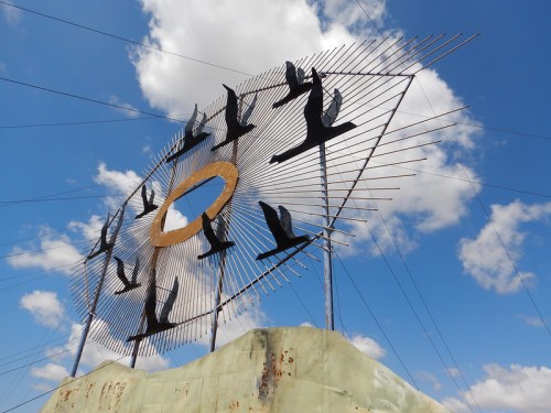 A large metal sculpture of birds in flight against a blue sky with clouds, featuring a circular golden element in the center.