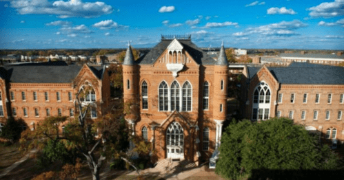 Historic brick building with gothic architecture, surrounded by trees and a blue sky with scattered clouds.