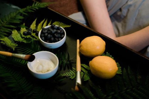 A tray with ferns, blueberries, two lemons, a small bowl of cream, and makeup brushes.
