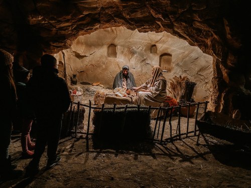 A dimly lit cave scene with figures seated, surrounded by straw and rustic decor, evoking a historical or biblical setting.