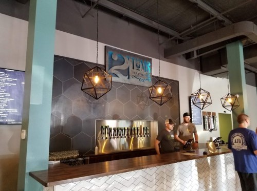 Interior of a brewery with a bar, two staff members, and geometric light fixtures. The brewery's logo is displayed prominently.