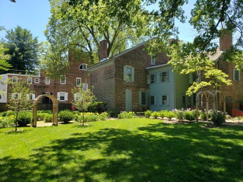 A sunny garden with lush green grass, featuring two historic stone buildings and blooming flowers.