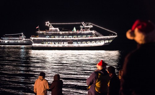 A decorated boat with lights sails on a dark water, while onlookers in winter attire watch from the shore.
