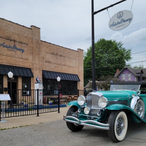 A vintage teal car parked outside a brewery with a brick facade and outdoor seating area. Green trees in the background.
