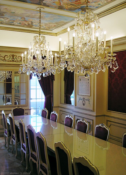Elegant dining room with a long table, ornate chandeliers, and decorative ceiling, featuring rich colors and luxurious furnishings.