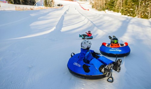 Three people tubing down a snowy slope, surrounded by trees and winter scenery.