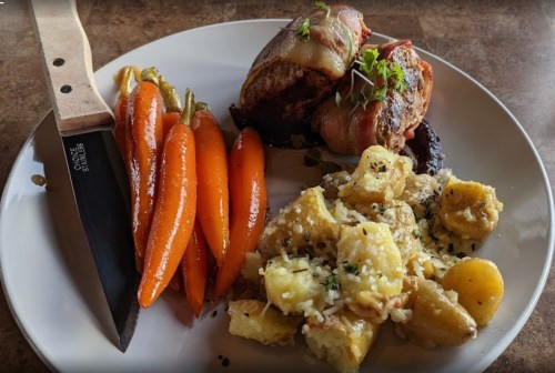 A plate with bacon-wrapped meat, roasted carrots, and cheesy potato gnocchi, accompanied by a knife.