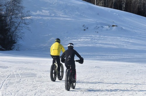 Two cyclists ride fat bikes on a snowy landscape, surrounded by trees and a snow-covered hill.
