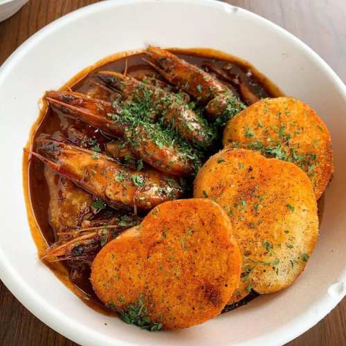 A bowl of shrimp in a rich sauce, garnished with herbs, accompanied by two golden, crispy bread pieces.