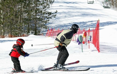 A child in a red helmet skis closely behind an adult on a snowy slope, with ski lifts and colorful markers in the background.
