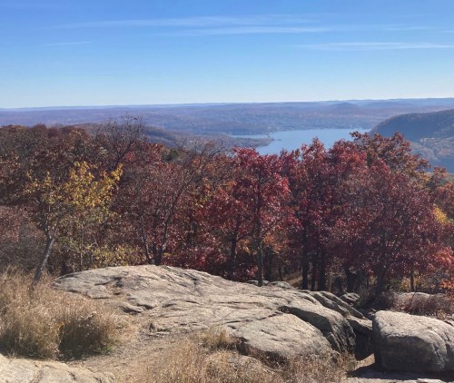 A scenic view from a rocky overlook, featuring vibrant autumn foliage and a lake in the distance under a clear blue sky.