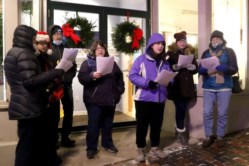 A group of people singing carols outside a shop, wearing winter clothing and holding song sheets, decorated for the holidays.