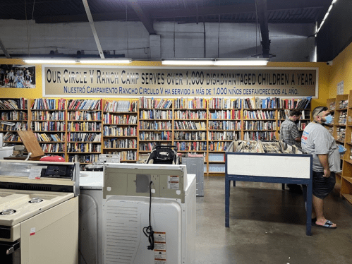 A spacious library with shelves of books and a sign about serving disadvantaged children. Two people are browsing.