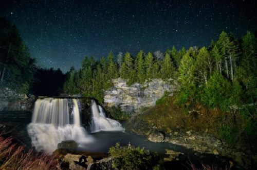 A serene waterfall cascades under a starry night sky, surrounded by lush trees and rocky cliffs.